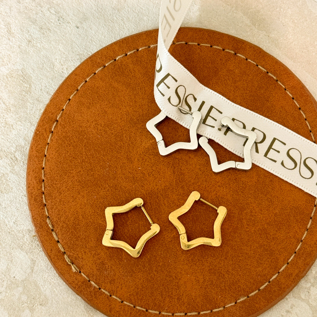 Gold star-shaped huggie earrings styled on brown suede jewelry pad with PRESSIE ribbon