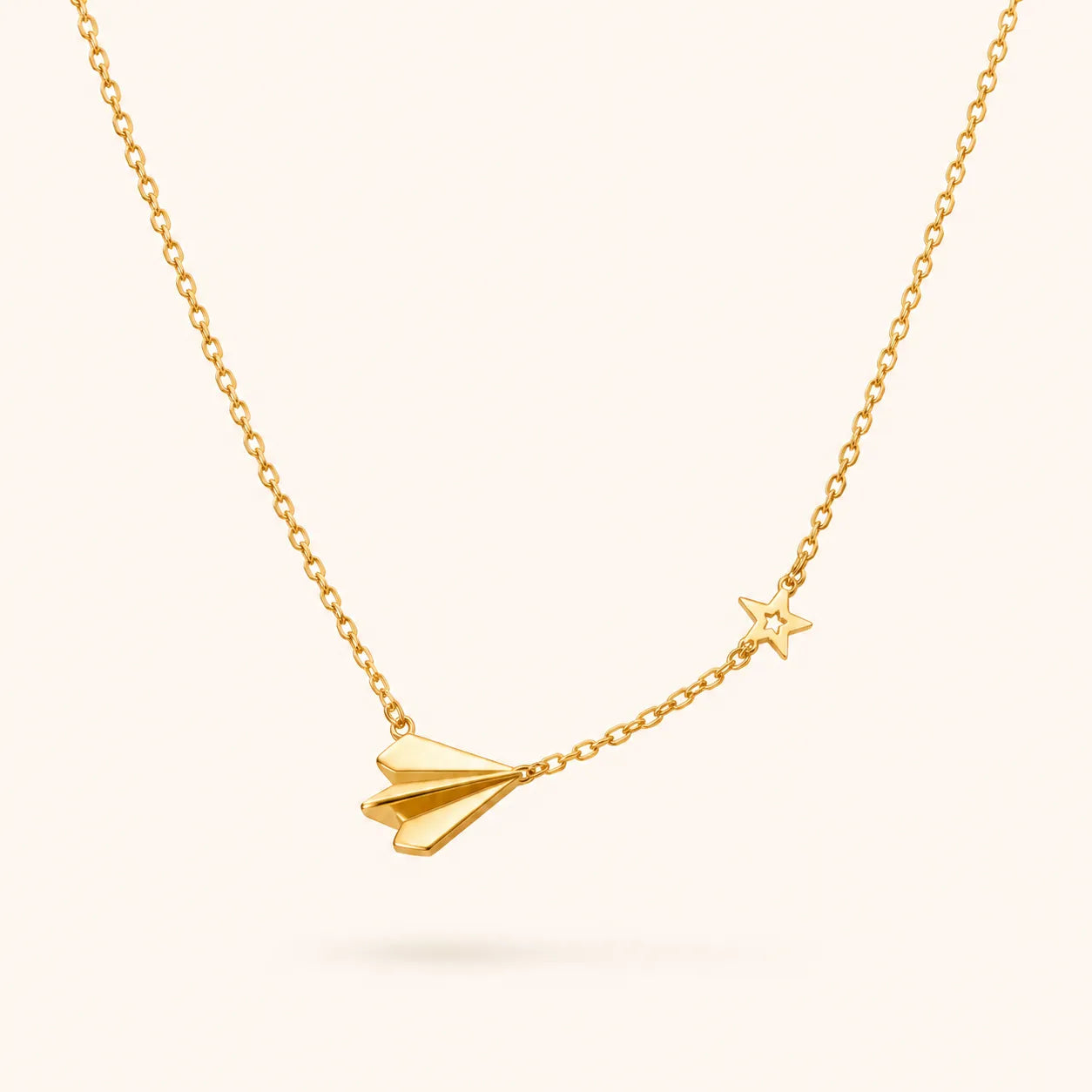 A delicate gold chain necklace featuring a small paper airplane charm and a tiny star charm, displayed against a solid pale pink background.