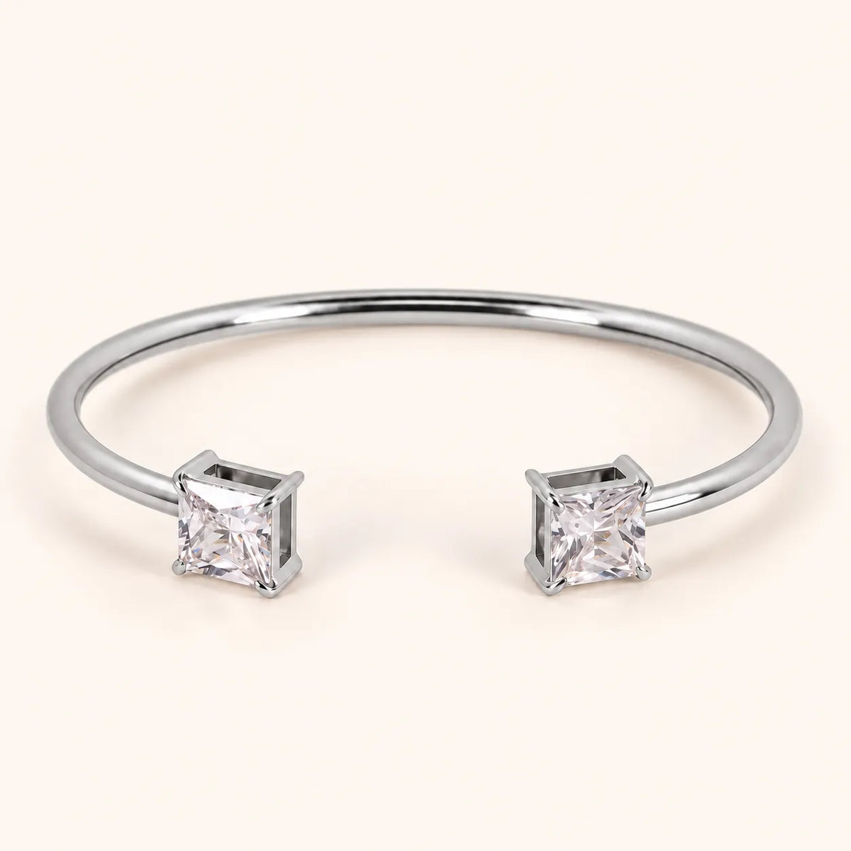 A silver-toned open cuff bangle bracelet with two princess-cut clear gemstones, shown on a light cream background.