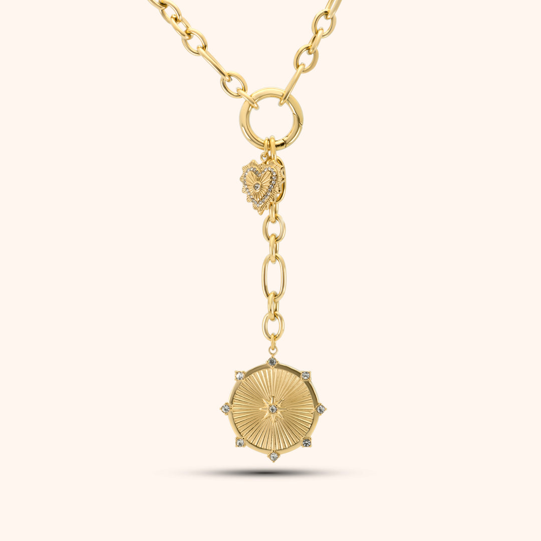 PRESSIE Radiant Heart Lariat necklace in 18k gold plate; features mixed paperclip chain, a central circular connector with a pavé heart charm, and a lariat drop ending in a sunburst medallion with clear stone accents.
