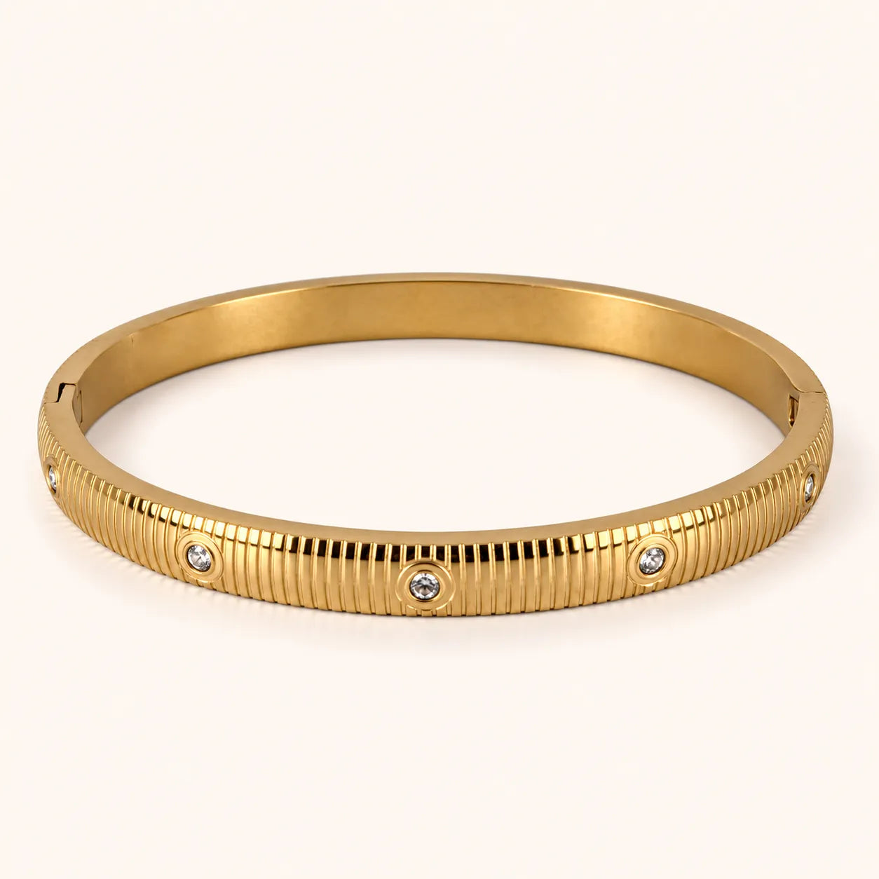 A gold bangle bracelet with a ribbed textured exterior and three small round clear stones set into the band, photographed on a clean off-white background.