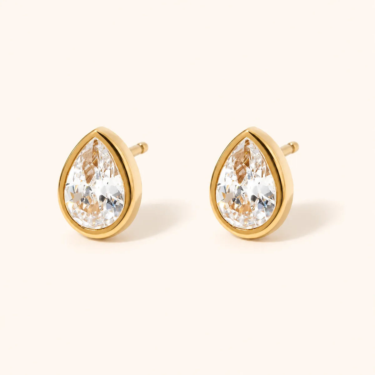 A pair of gold teardrop stud earrings with clear faceted gemstones, displayed on a light cream background.