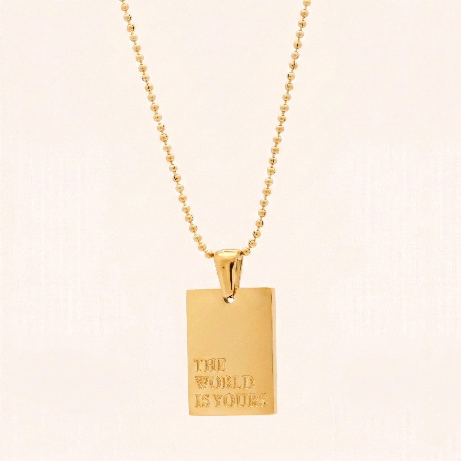 Gold necklace with a pendant that says 'The World is Yours' on a beige background