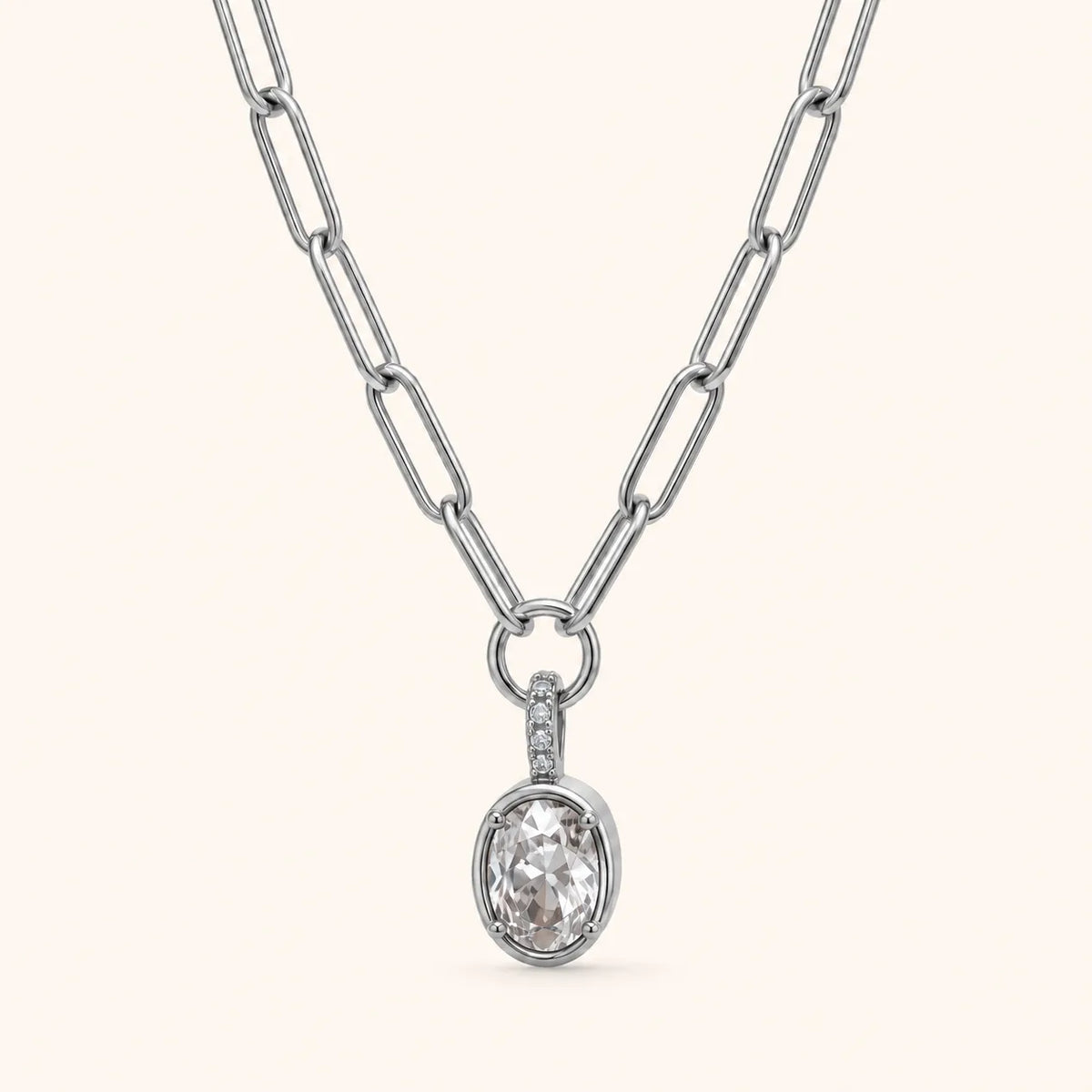 A silver paperclip chain necklace with an oval pendant featuring a clear faceted gemstone and a small side accent, shown on a light cream background.