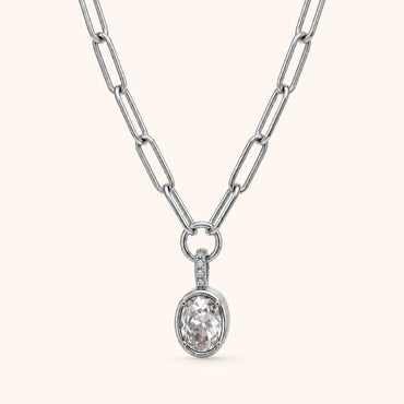 A silver paperclip chain necklace with an oval pendant featuring a clear faceted gemstone and a small side accent, shown on a light cream background.