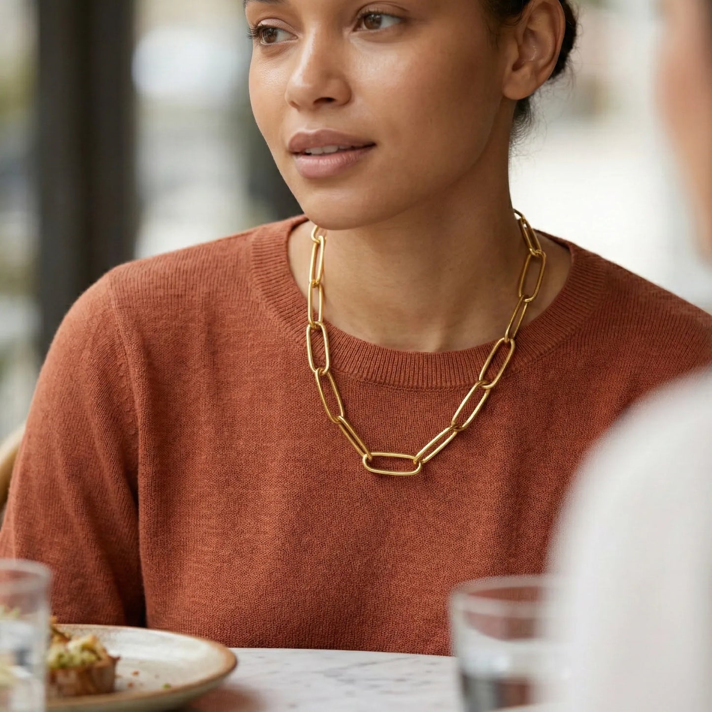 Woman wearing a gold chain necklace sitting at a table with a blurred background