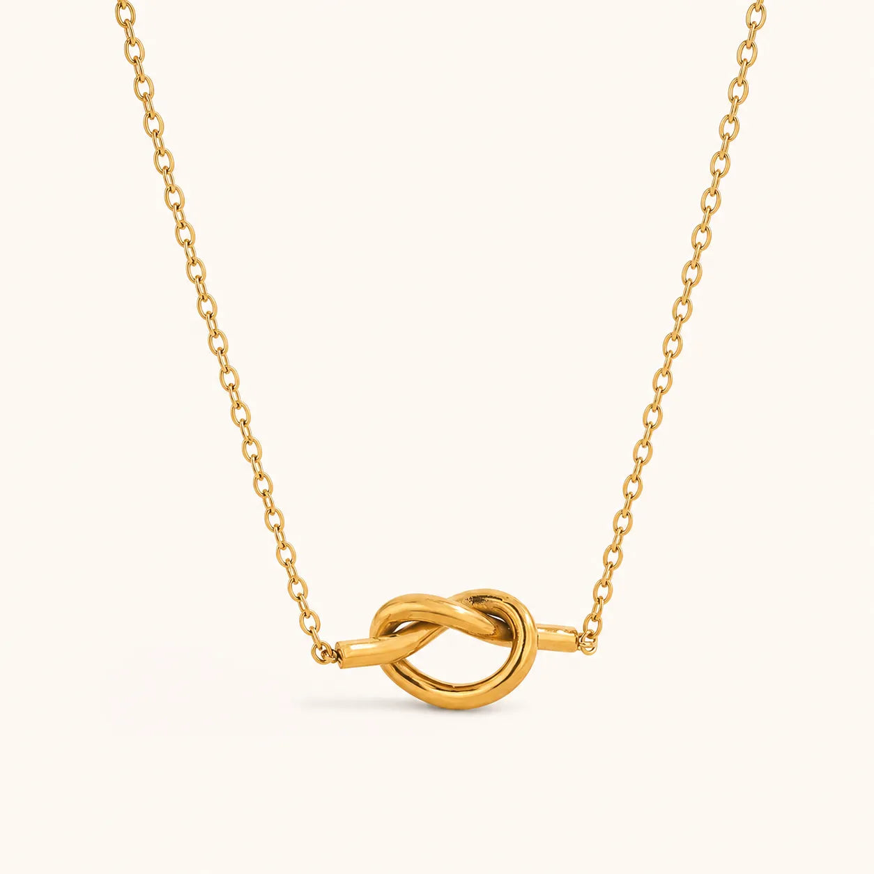 A gold chain necklace with a polished knot pendant, centered on a light cream background.