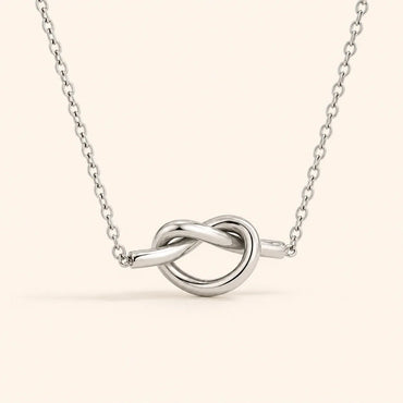 A silver necklace with a polished knot pendant, suspended from a delicate link chain against a solid pale beige background.