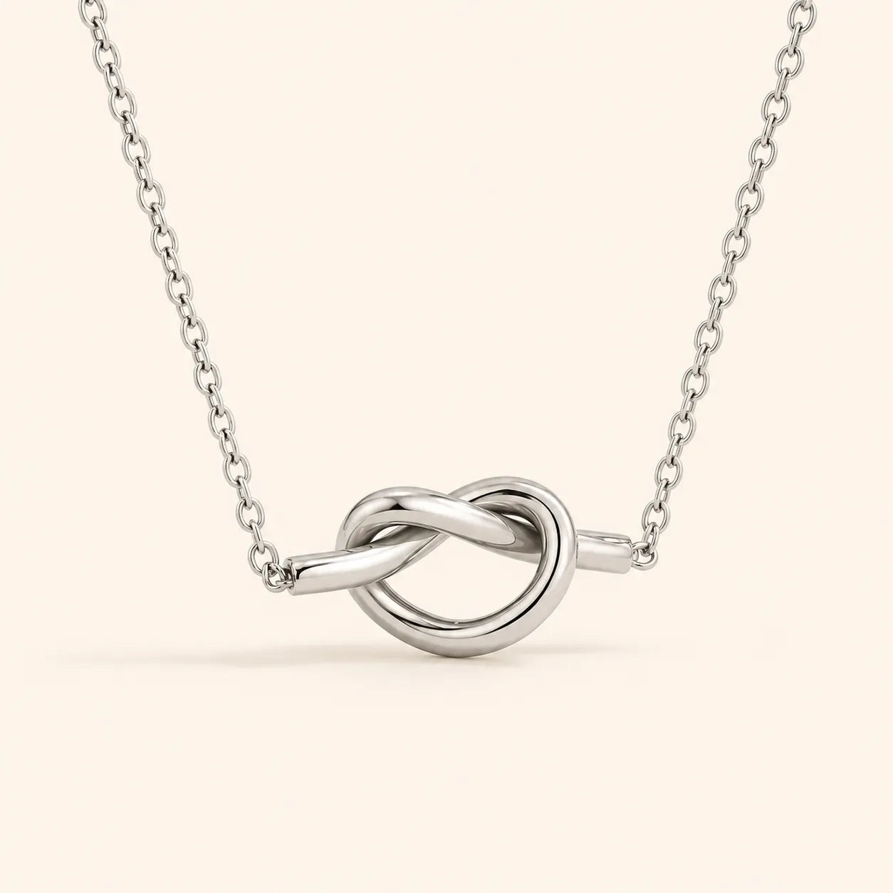 A silver necklace with a polished knot pendant, suspended from a delicate link chain against a solid pale beige background.
