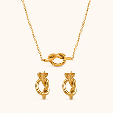 Gold jewelry set featuring a delicate chain necklace with a central knotted loop pendant and matching stud earrings with circular and knot details, displayed on a clean off-white background.