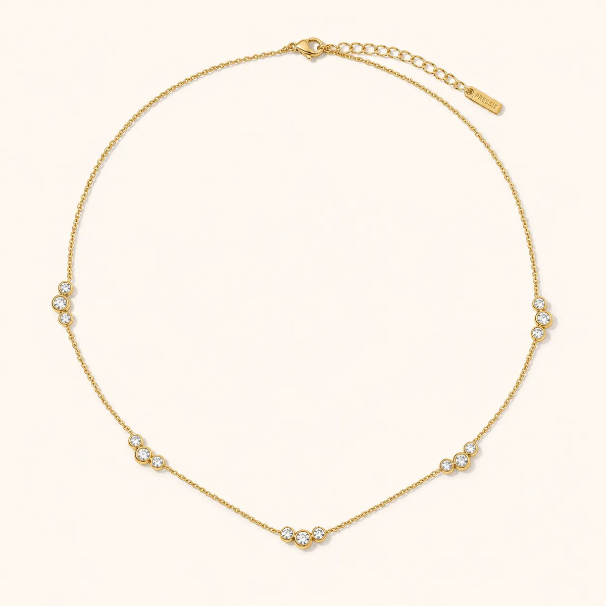 A delicate gold chain necklace with five small clusters of clear round stones, arranged in a circle on a warm off-white background. The brand name 'SARAH' is visible on a small gold tag near the clasp.