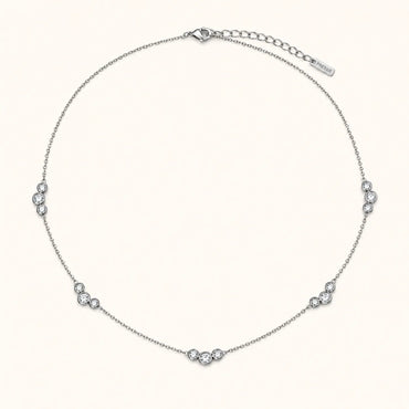 A delicate silver chain necklace with five small clusters of clear round-cut stones spaced evenly along the band, displayed on a light cream background. A brand tag is visible on the extender chain.