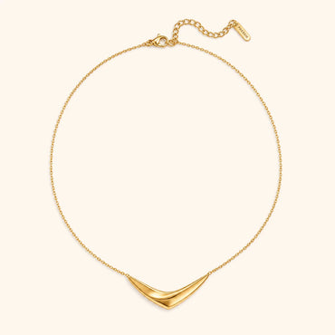 A delicate gold chain necklace with a wide, curved chevron pendant, displayed on a light cream background.