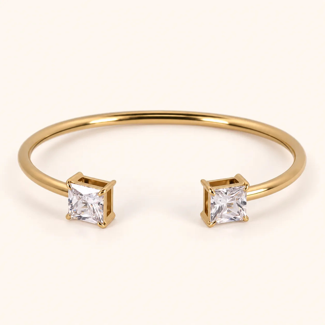 A gold open cuff bracelet featuring two princess-cut clear gemstones in square bezel settings, shown on a light cream background.