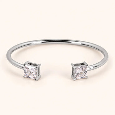 A silver-toned open cuff bangle bracelet with two princess-cut clear gemstones, shown on a light cream background.