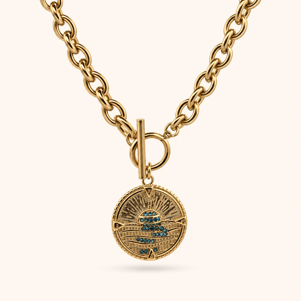 A gold chain necklace with a round medallion pendant featuring a stylized sun and moon design accented with small blue stones, shown against a light cream background.