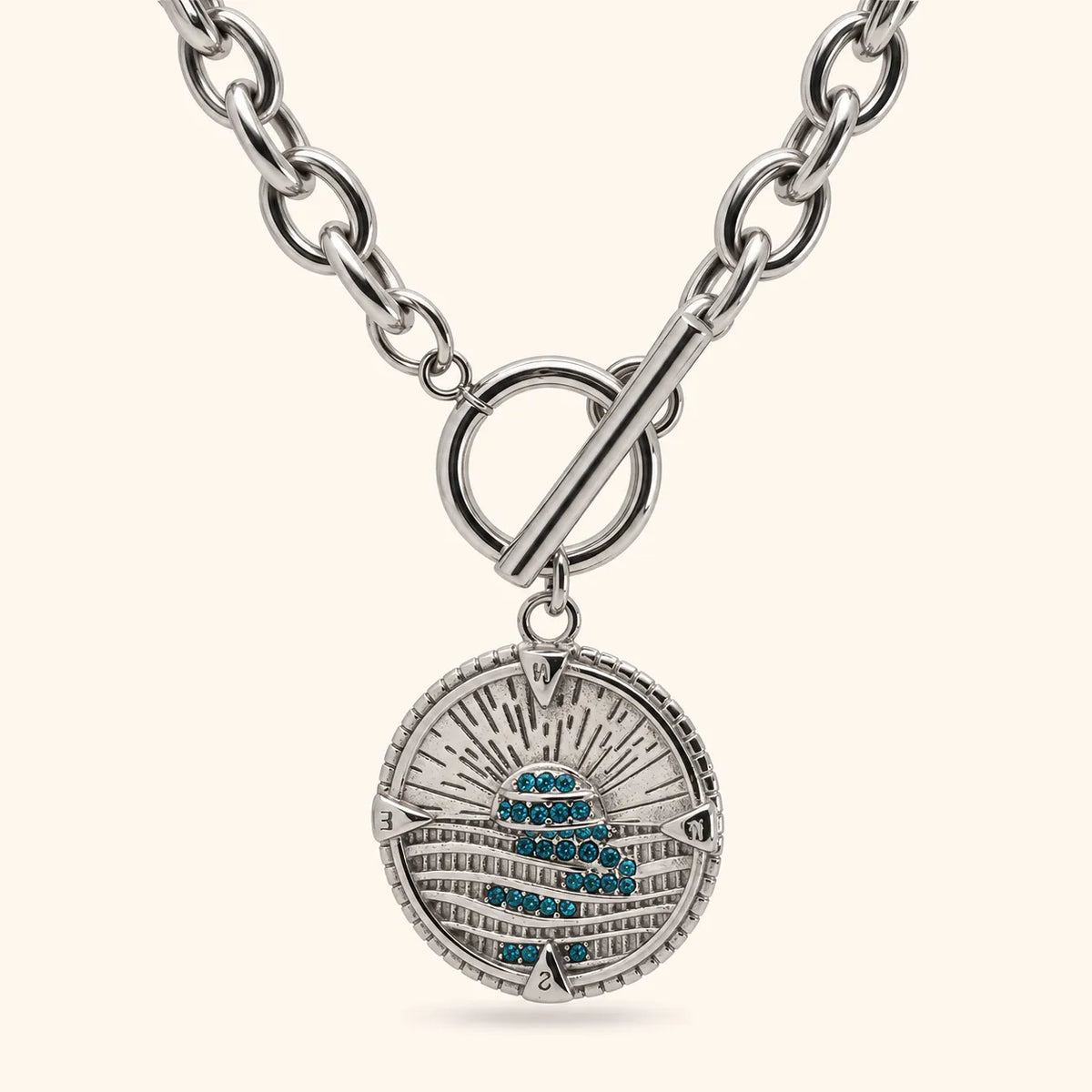 A silver-toned round pendant necklace with a toggle clasp, featuring a textured sunburst design and a central cluster of small blue stones, shown on a light cream background.