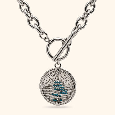 A silver-toned round pendant necklace with a toggle clasp, featuring a textured sunburst design and a central cluster of small blue stones, shown on a light cream background.
