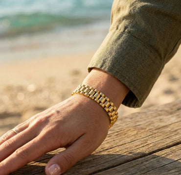 Hand wearing a gold bracelet on a wooden surface with a beach background